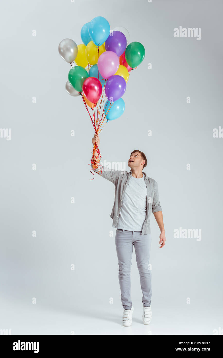 Schöner Mann stehen auf die Zehenspitzen und halten Bündel von bunten Luftballons auf grauem Hintergrund Stockfoto