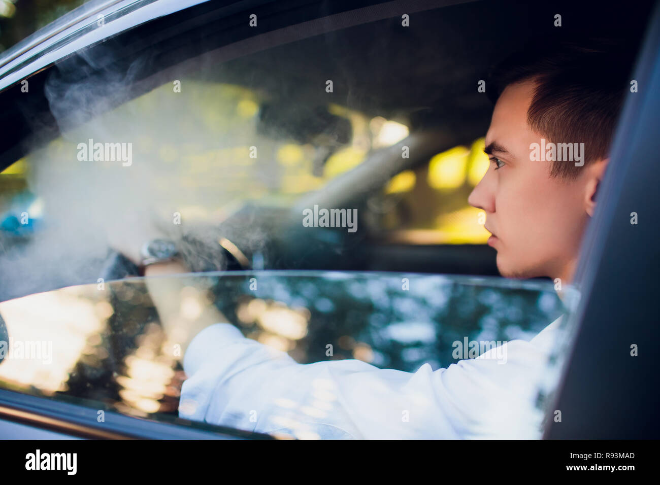 elfbar elfa master akku Ansicht von der Seite junge Mann Rauchen einer E-Zigarette wie er fährt mit  seinem Auto auf einer städtischen Straße. Autofahrer lugt aus dem Auto und  raucht Stockfotografie