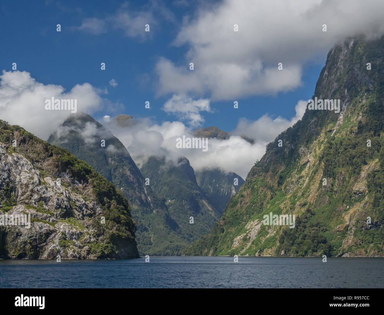 Blue Sky gibt Weg für flauschige Wolken an den Spitzen von steilen Bergen aus dem Fjord Gewässern Neuseelands Fiordlands Nationalpark steigende Stockfoto