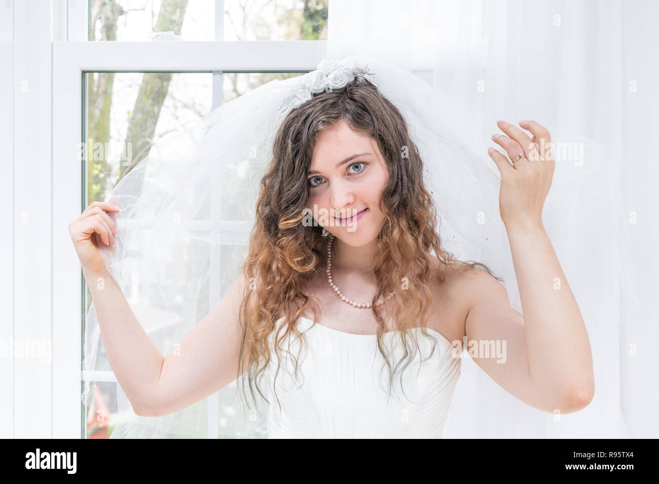 Closeup Portrait von jungen weiblichen Person, Frau, Braut im Brautkleid, Schleier, Hände, Gesicht, Perlenkette, Fenster, weiße Vorhänge, Stockfoto