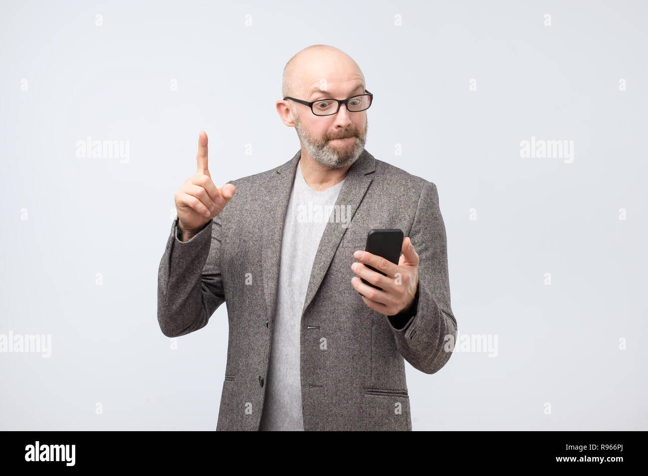 Mann hebt Zeigefinger, blickt mit großem Interesse an Smart Phone, liest relevante Nachrichten im Internet Stockfoto