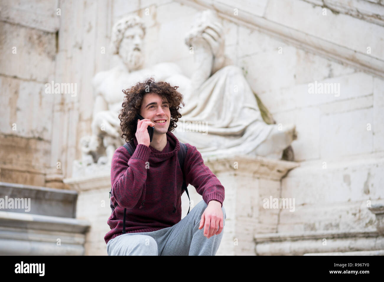 Hübscher junger Mann mit lockigem Haar im Trainingsanzug über sein Handy vor Nil Gott Statue in Rom Stockfoto
