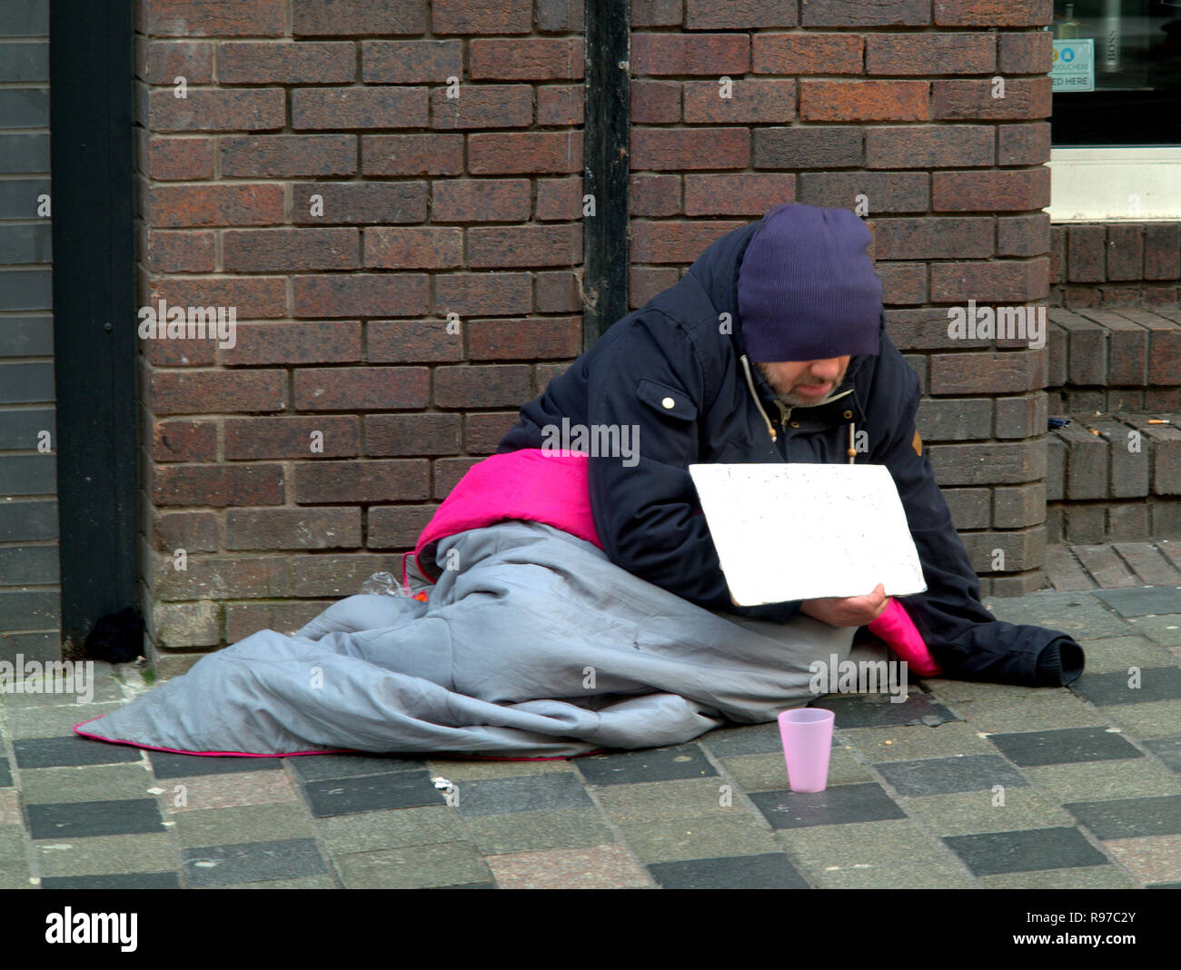 Obdachloser betteln auf der Straße mit Ziegel Wand rosa Schale verbergen Gesicht halten Schild mit Schlafsack Stockfoto