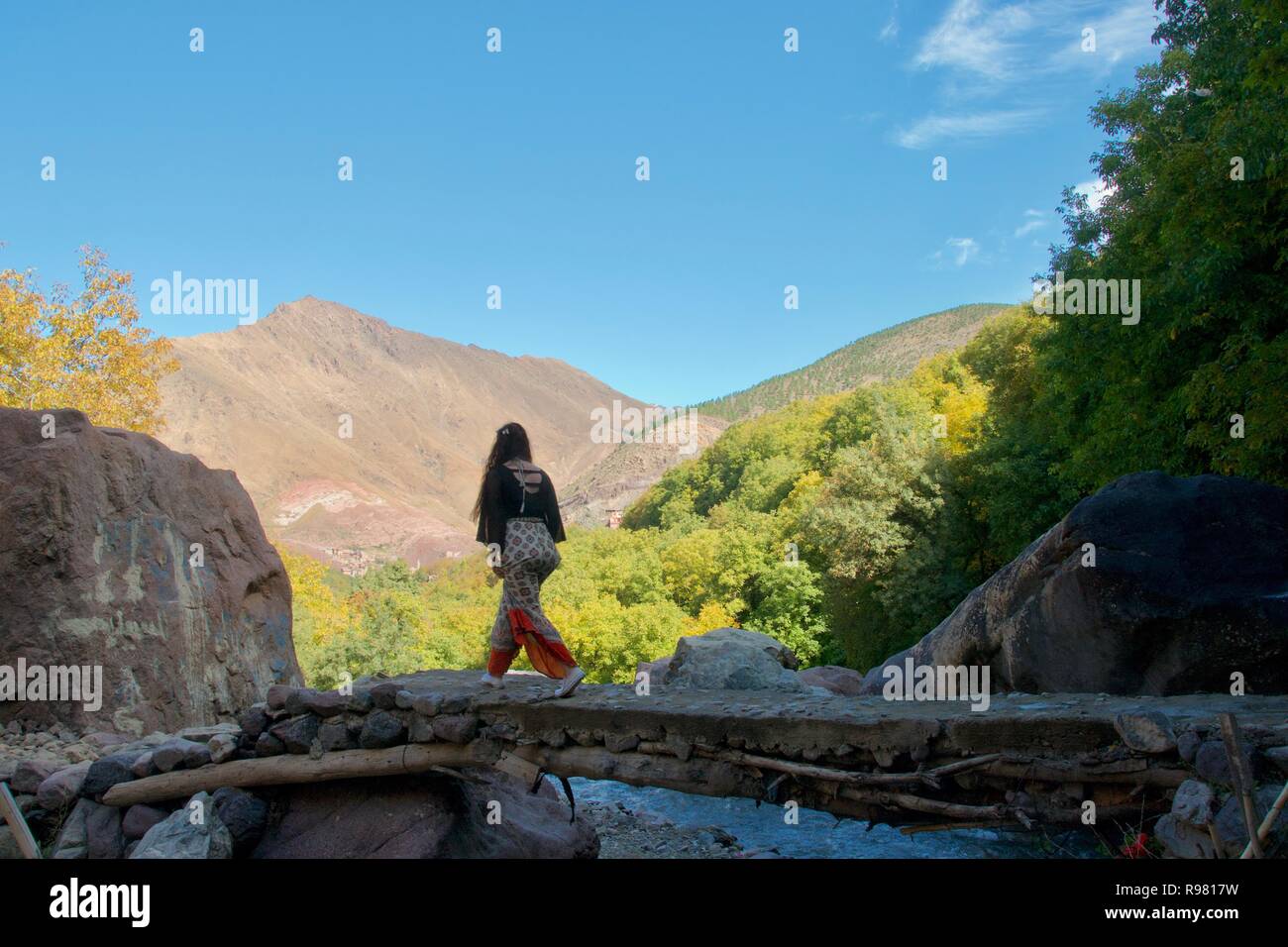 Eine weibliche Touristen überquert eine Brücke über einen Bach in den Ausläufern des Atlasgebirges. Die bewaldeten Imlil Tal und die Berge sind hell erleuchtet in der Stockfoto