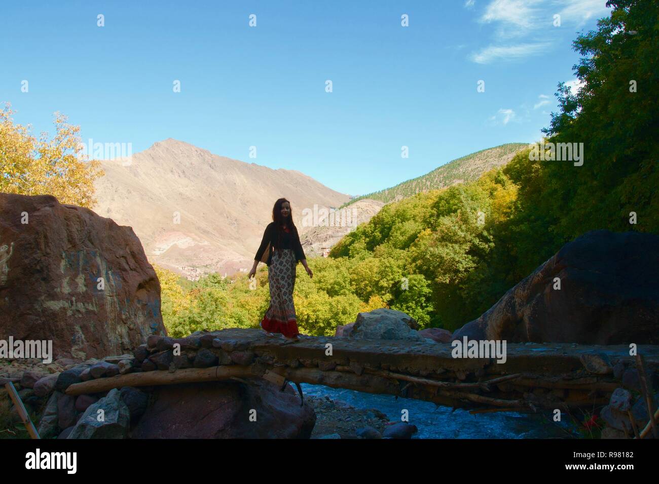 Eine weibliche Touristen kreuzt einen schattigen Brücke über einen Bach in den Ausläufern des Atlasgebirges. Die bewaldeten Imlil Tal und die Berge sind hell erleuchtet Stockfoto
