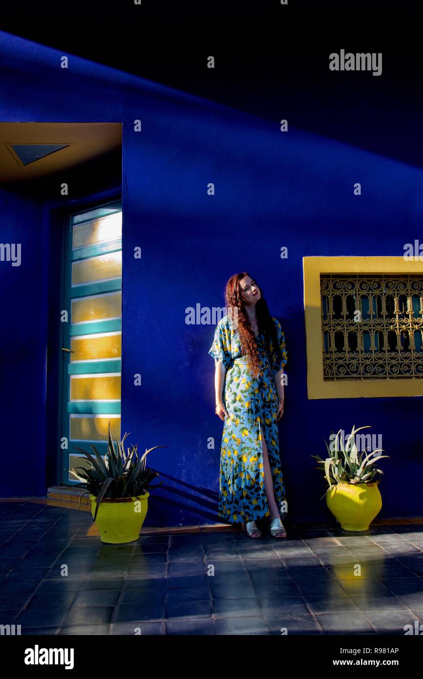 Eine langhaarige Brünette Dame trägt einen blauen und gelben Zitrone gemusterten Kleid sieht nach oben, auf eine blaue und gelbe Garten Terrasse posiert, beleuchteten seitlich durch Stockfoto