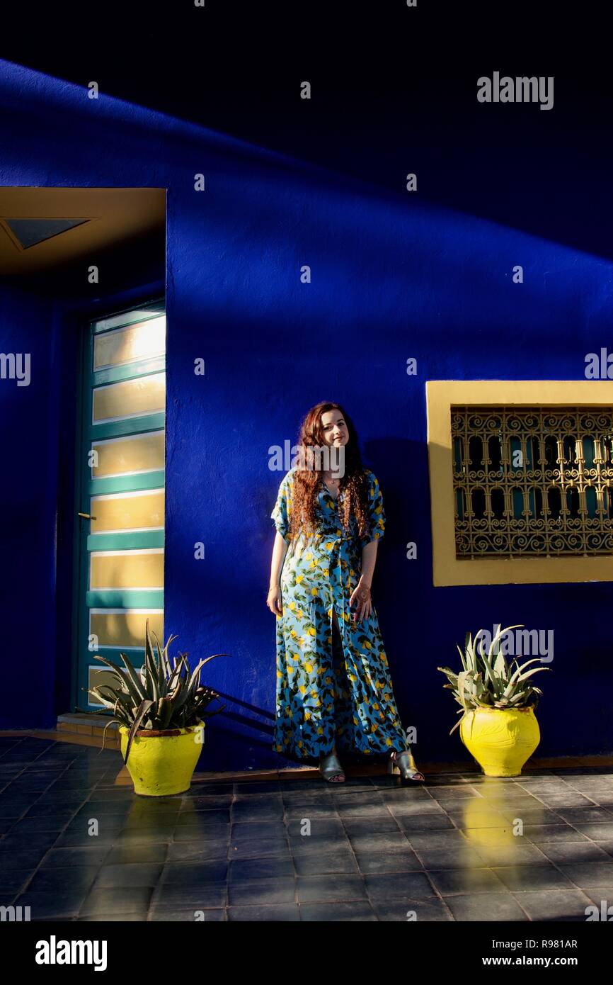 Eine langhaarige Brünette Dame trägt einen blauen und gelben Zitrone gemusterten Kleid auf einem blauen und gelben Garten Terrasse Posen, beleuchteten seitlich durch helles Sonnenlicht Stockfoto