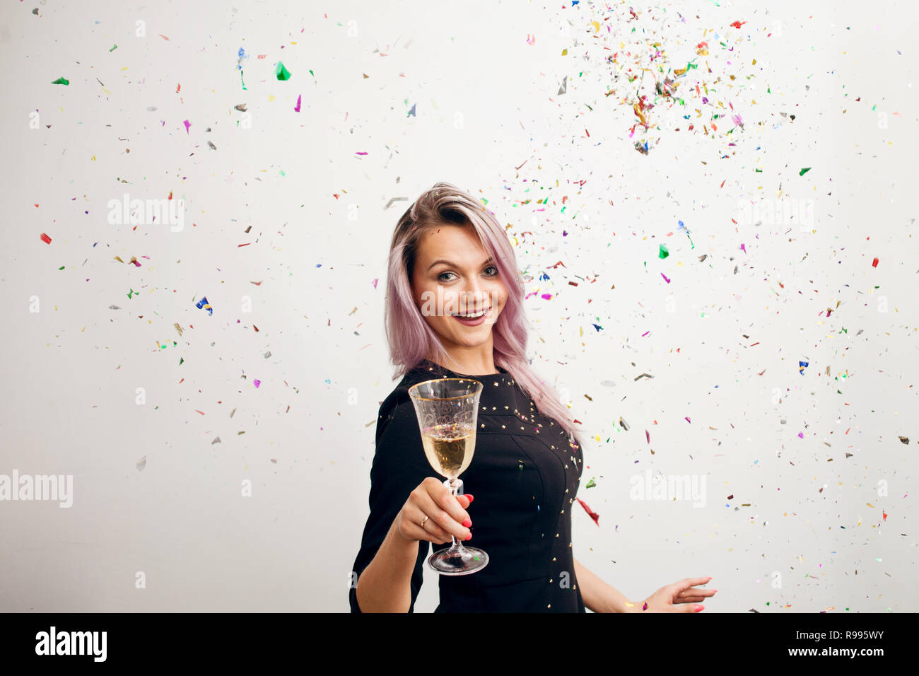 Lustige junge Frau in schwarzem Kleid zu feiern. Schöne junge Frau mit einem Glas Champagner und Konfetti Stockfoto