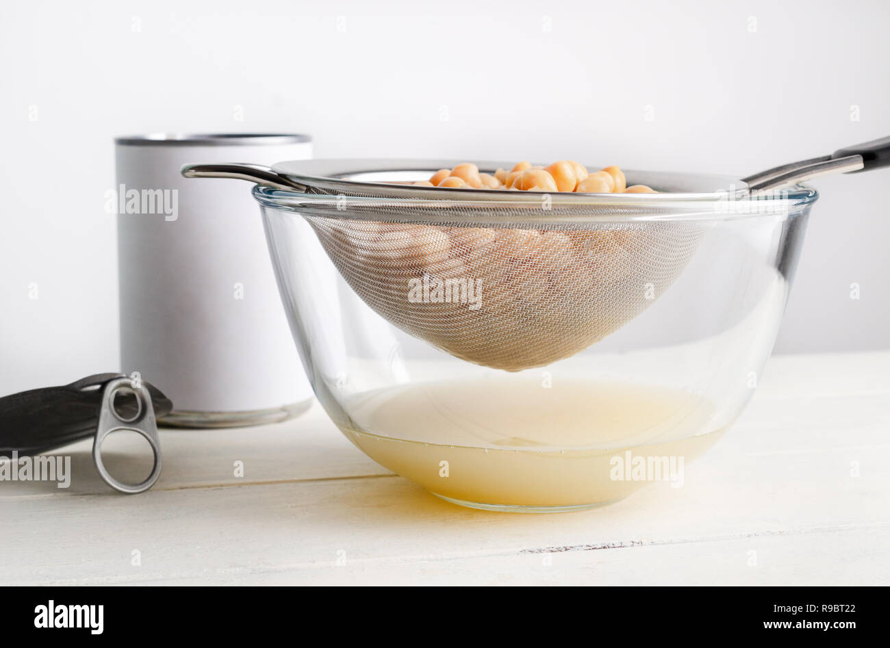 Metallsieb entleeren aquafaba Kichererbse Wasser in Glasschale. Unbranded geöffnete Dose mit Deckel ruht auf Laufbelag Tabelle links. Augenhöhe erschossen. Stockfoto