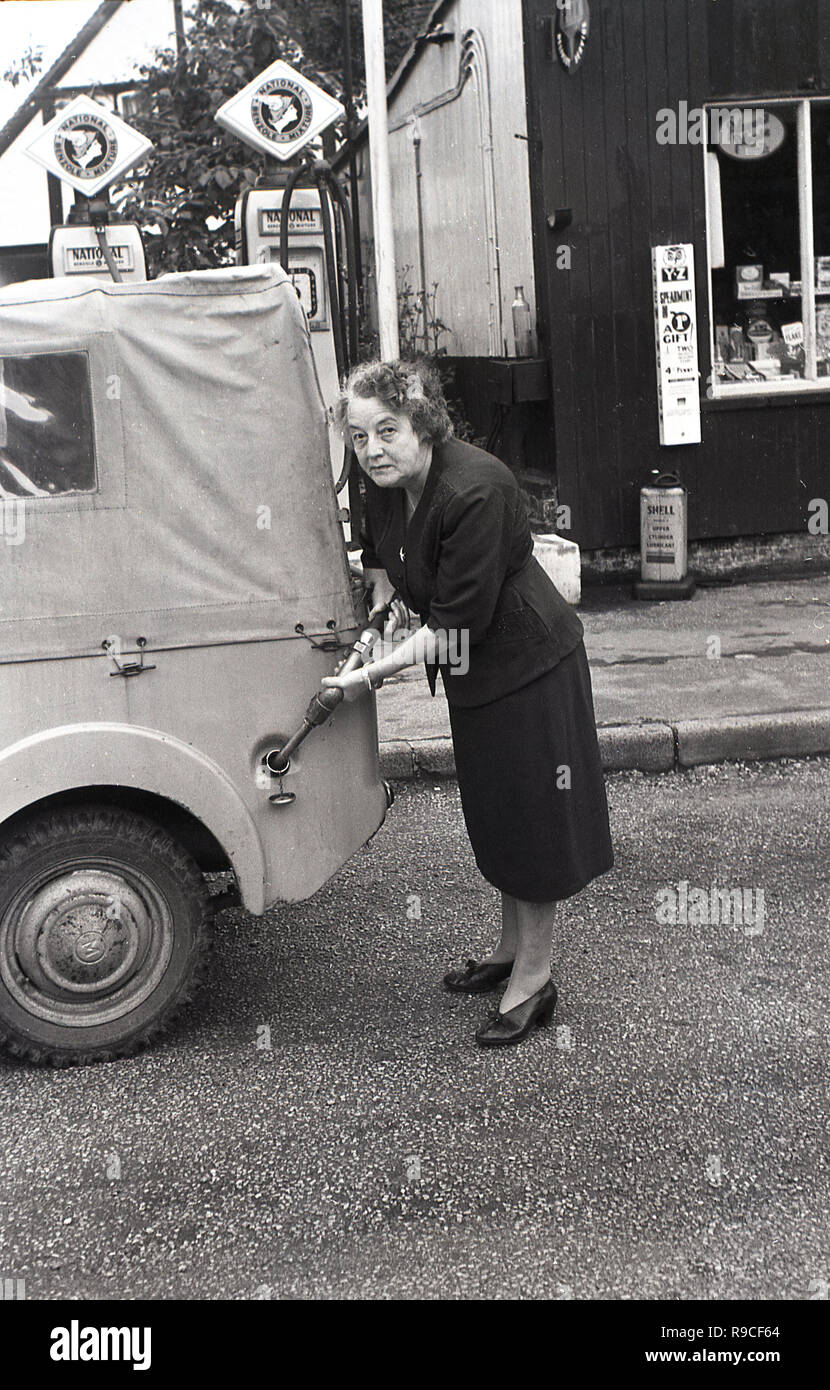 1955, historische, in einem ländlichen High Street, eine ältere Frau mit Benzin pumpe mit Kraftstoff füllen, Ihre Leinwand überdachten kleinen Jeep oder Kraftfahrzeug, England, UK. Stockfoto