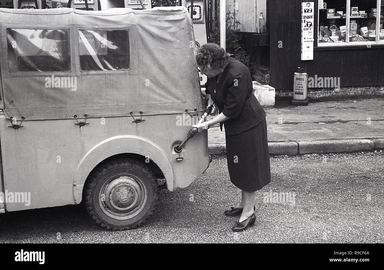 1955, historische, in einem ländlichen High Street, eine Frau mit Benzin pumpe mit Kraftstoff füllen, Ihre Leinwand überdachten kleinen Jeep oder Kraftfahrzeug, England, UK. Stockfoto