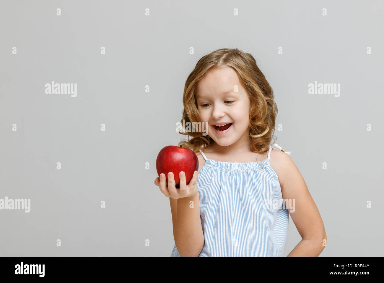 Porträt einer Glücklich lächelnde kleine blonde Mädchen auf einem grauen Hintergrund. Das Kind ist auf der Suche und bereit, roten Apfel zu essen Stockfoto