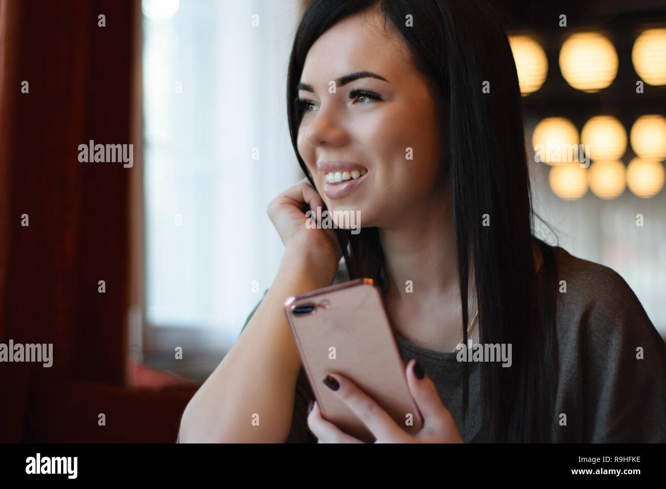 Eine lächelnde schöne Mädchen mit Grübchen auf den Wangen in einem gemütlichen Cafe gegen das Fenster und Bokeh. attraktives Modell das Telefon hält Stockfoto