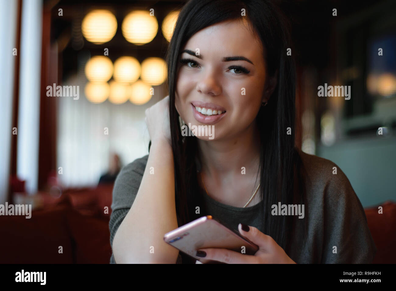 Eine lächelnde schöne Mädchen mit Grübchen auf den Wangen in einem gemütlichen Cafe gegen das Fenster und Bokeh. attraktives Modell das Telefon hält Stockfoto