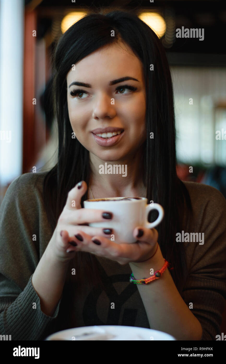 Schöne Mädchen mit Grübchen auf den Wangen in einem gemütlichen Cafe gegen das Fenster und Bokeh. attraktive Modell mit der Tasse Kaffee schaut aus dem Fenster Stockfoto