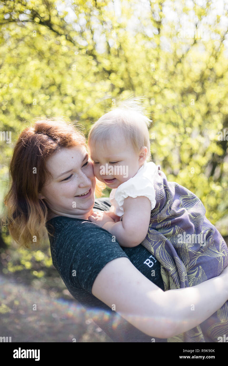Mutter lächelnd, während sie ihr Baby wickeln in einer Schlinge für babywearing. Lifestyle Bild geschossen auf Lage in einem Park an einem sonnigen Frühlingstag. Stockfoto