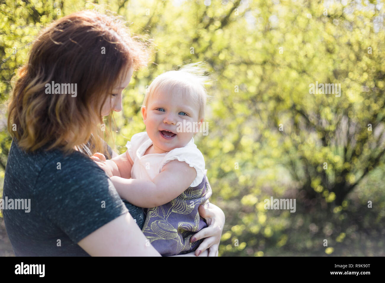 Mutter lächelnd, während sie ihr Baby wickeln in einer Schlinge für babywearing. Lifestyle Bild geschossen auf Lage in einem Park an einem sonnigen Frühlingstag. Stockfoto