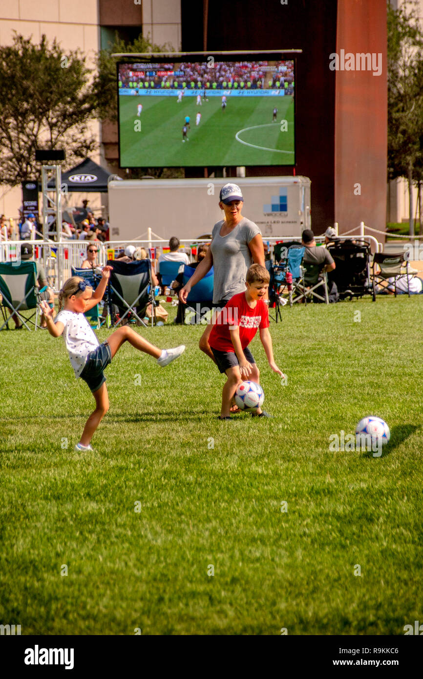 Während tagsüber ein Publikum Uhren eine Fußball-WM Spiel auf einen gigantischen Outdoor TV-Bildschirm in Costa Mesa, CA, eine Mutter mit ihren Kindern das Spiel auf einem nahe gelegenen Feld spielt. Stockfoto