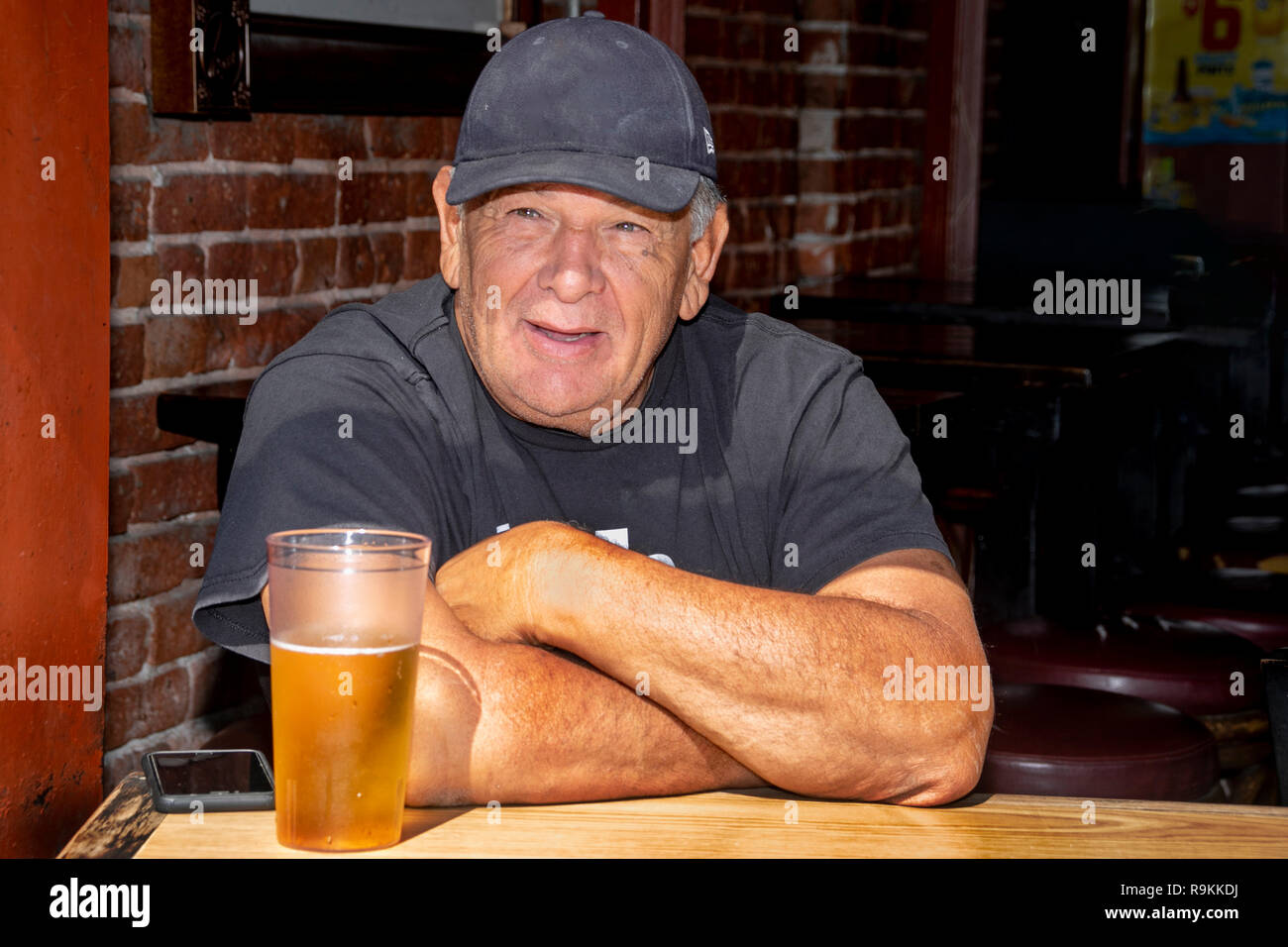 Ein herzhaftes älterer Mann in Schwarz Kappe und T-Shirt genießt ein frisch gezapftes Bier im Fenster eines Huntington Beach, CA, Taverne an einem warmen Sommermorgen. Stockfoto