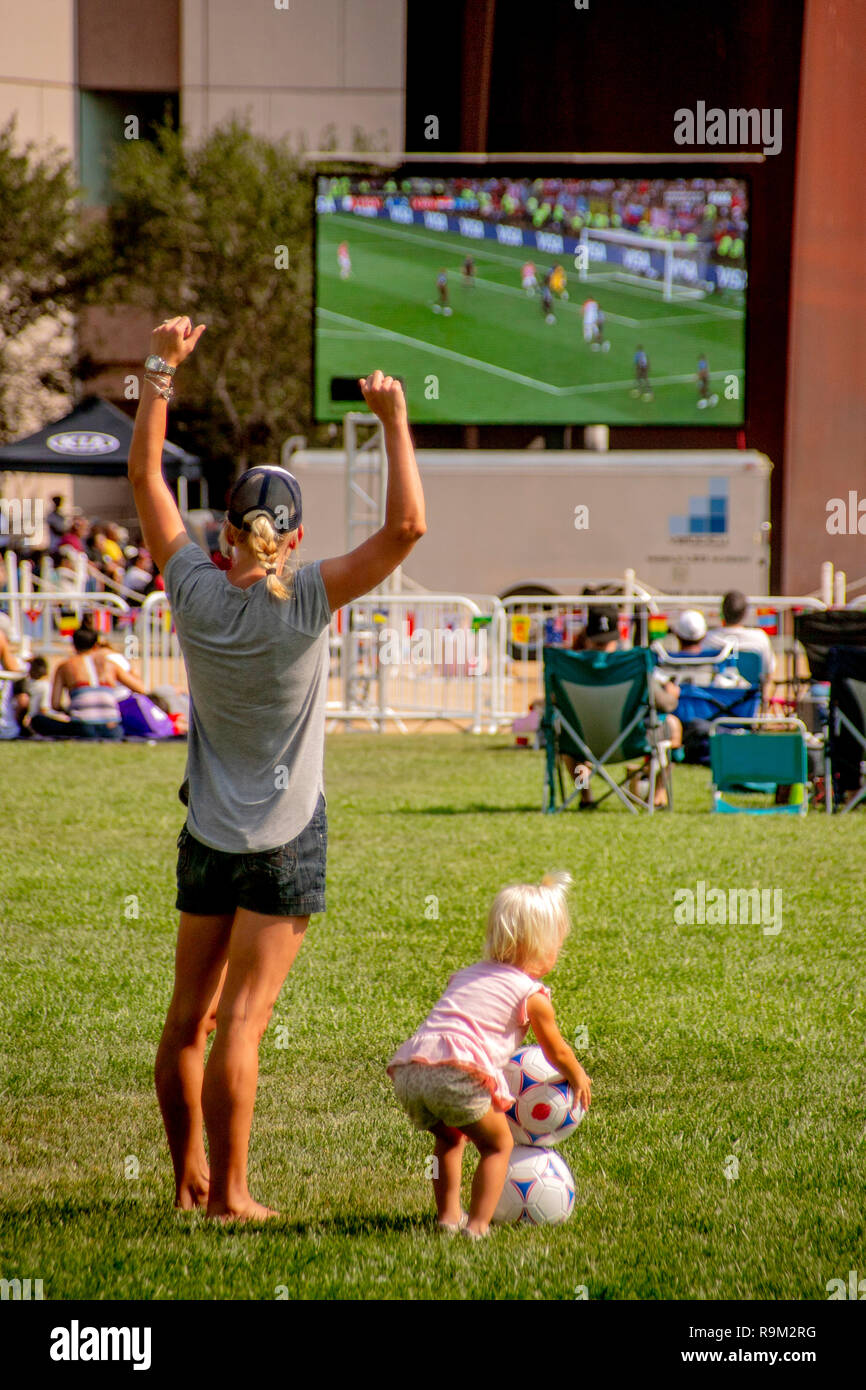 Während tagsüber ein Publikum Uhren eine Fußball-WM Spiel auf einen gigantischen Outdoor TV-Bildschirm in Costa Mesa, CA, eine Mutter mit ihren Kindern das Spiel auf einem nahe gelegenen Feld spielt. Stockfoto