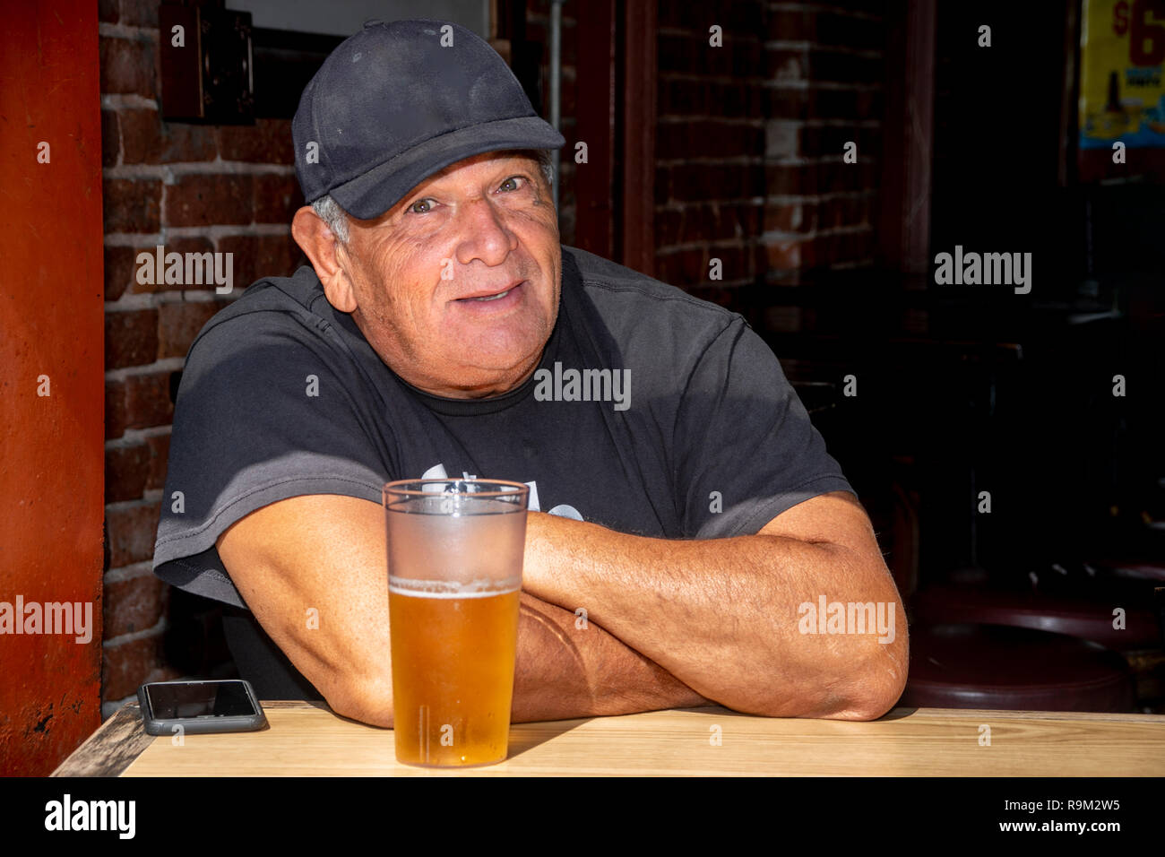 Ein herzhaftes älterer Mann in Schwarz Kappe und T-Shirt genießt ein frisch gezapftes Bier im Fenster eines Huntington Beach, CA, Taverne an einem warmen Sommermorgen. Stockfoto