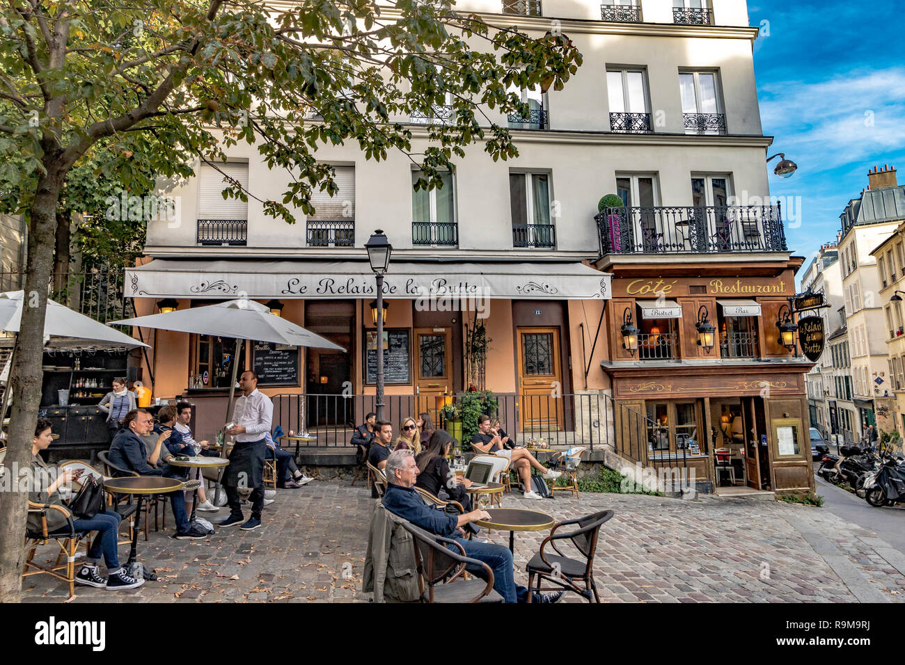 Leute, die sich an Tischen vor dem Restaurant Le Relais de la butte entspannen, während ein Kellner eine Bestellung eines Kunden in Montmartre, Paris, annimmt Stockfoto