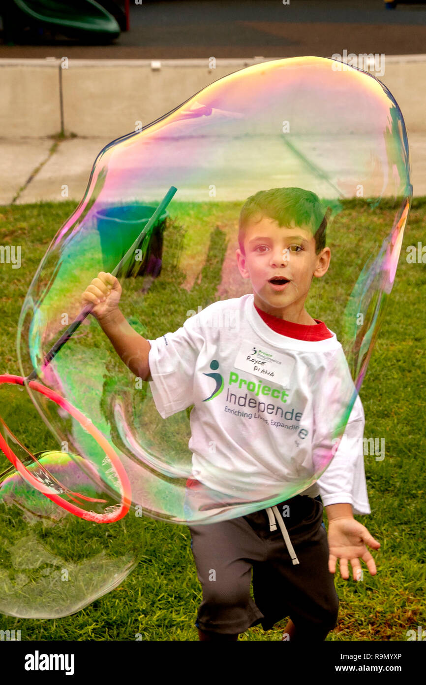 Zu einem Charity März für Behinderte in Costa Mesa, CA, ein Hispanic junge ist fasziniert, Während weht eine ornamentale Seifenblase. Hinweis Logo T-Shirt. Stockfoto