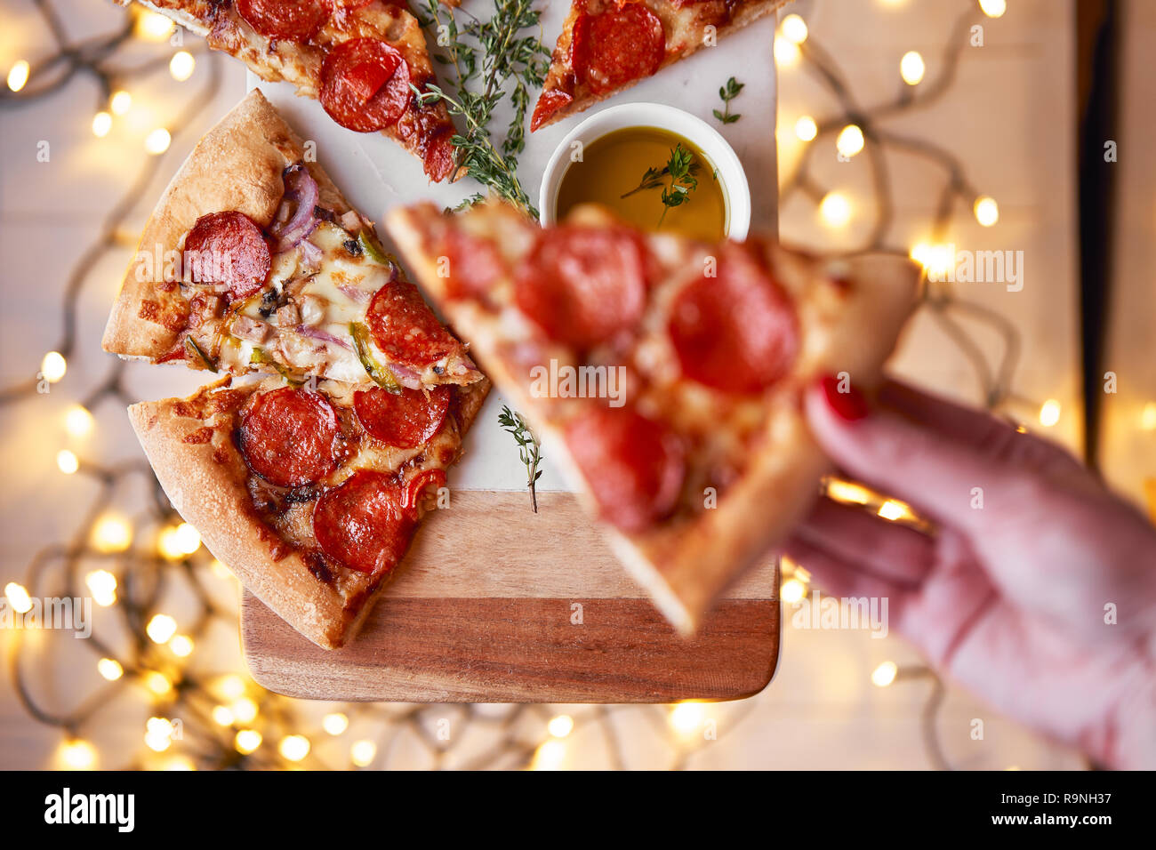 Weihnachten und Neujahr Atmosphäre. Womans hand nimmt Scheibe der Italienischen Pizza mit Schmelzen mit Tomate, Salami und Käse auf einem weißen Marmor Schneidebrett. Stockfoto