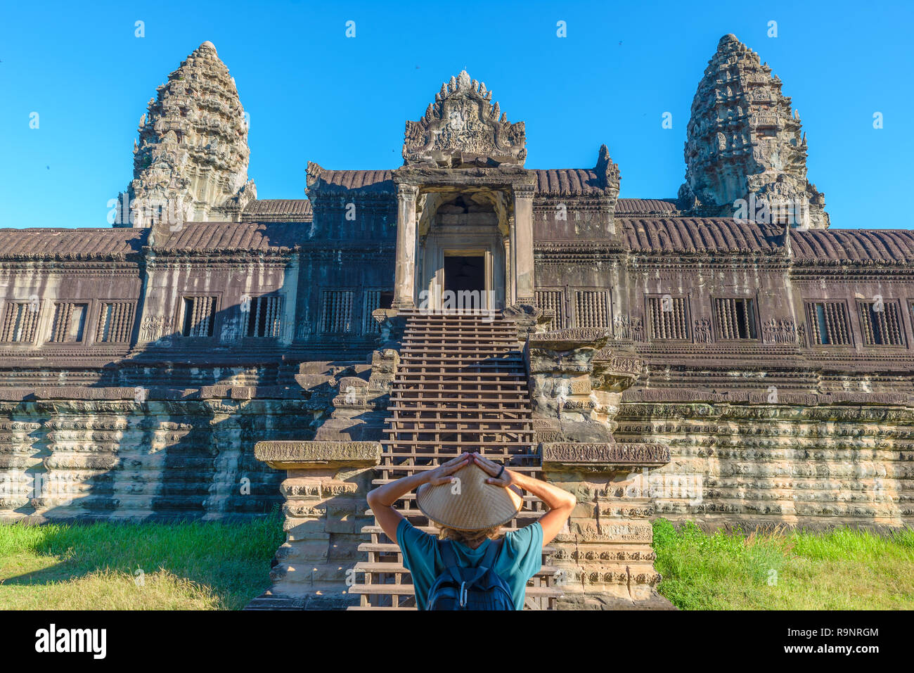 Eine touristische besuchen die Ruinen von Angkor Wat bei Sonnenaufgang, Reiseziel Kambodscha. Frau mit traditionellen Hut und erhobenen Armen, Rückansicht, Hauptfassade stai Stockfoto