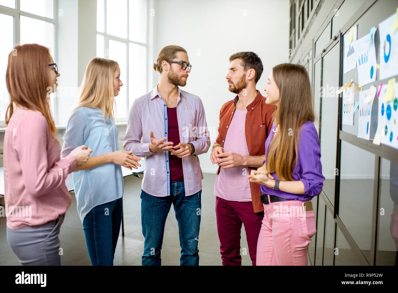 Junges Team von Mitarbeitern gekleidet beiläufig zusammen gesprochen in der Nähe der Glaswand mit einige Statistiken und Aufkleber im Büro Stockfoto