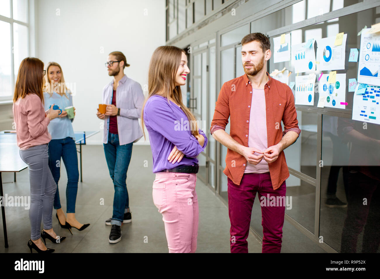 Junges Team von Mitarbeitern gekleidet beiläufig im Gespräch in der Nähe der Glaswand mit einige Statistiken und Aufkleber im Büro Stockfoto