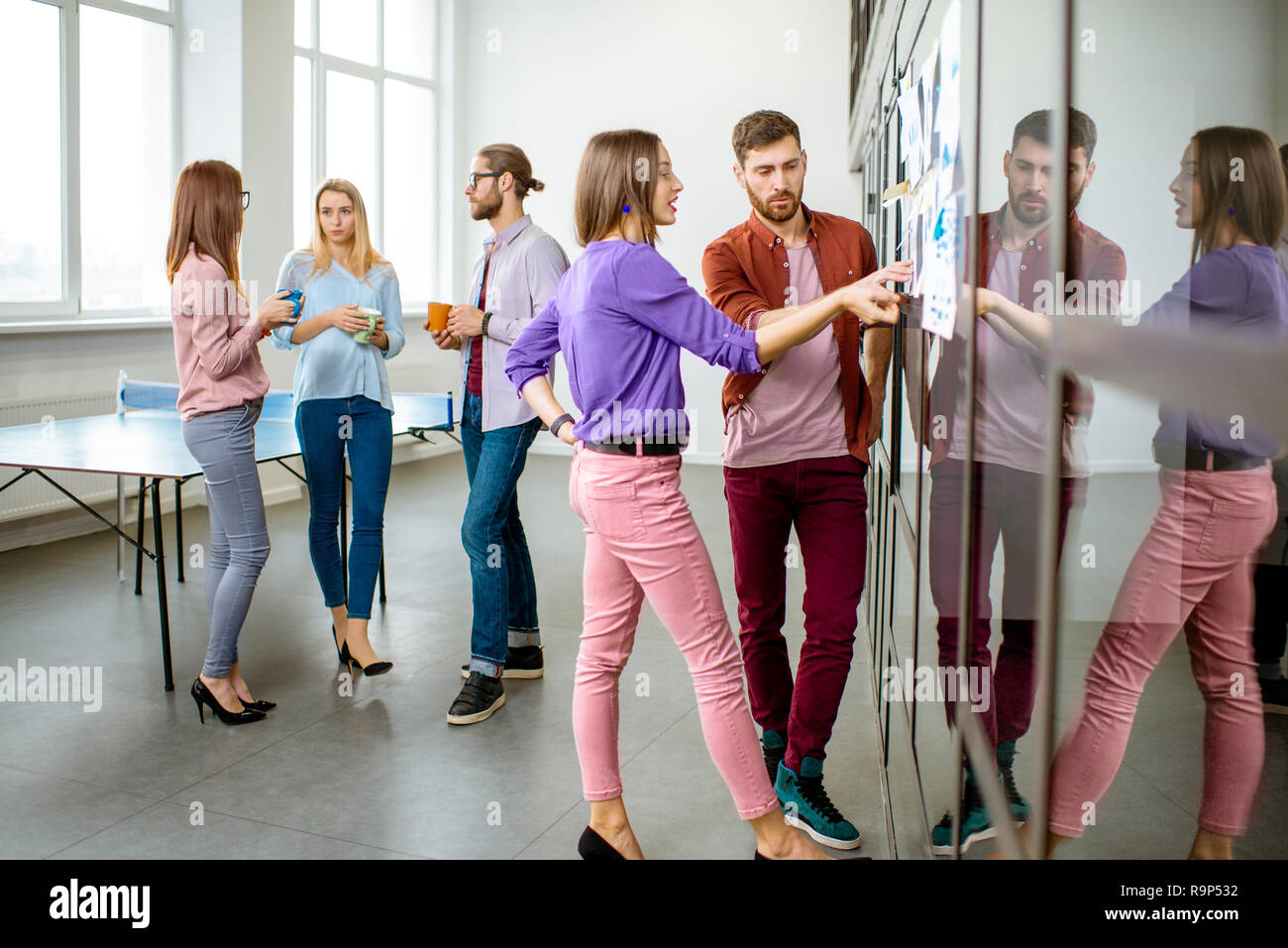 Junges Team von Mitarbeitern gekleidet beiläufig im Gespräch in der Nähe der Glaswand mit einige Statistiken und Aufkleber im Büro Stockfoto