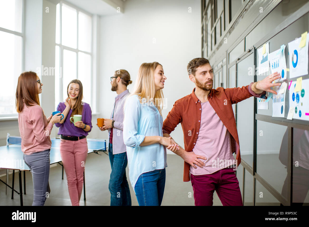 Junges Team von Mitarbeitern gekleidet beiläufig zusammen gesprochen in der Nähe der Glaswand mit einige Statistiken und Aufkleber im Büro Stockfoto