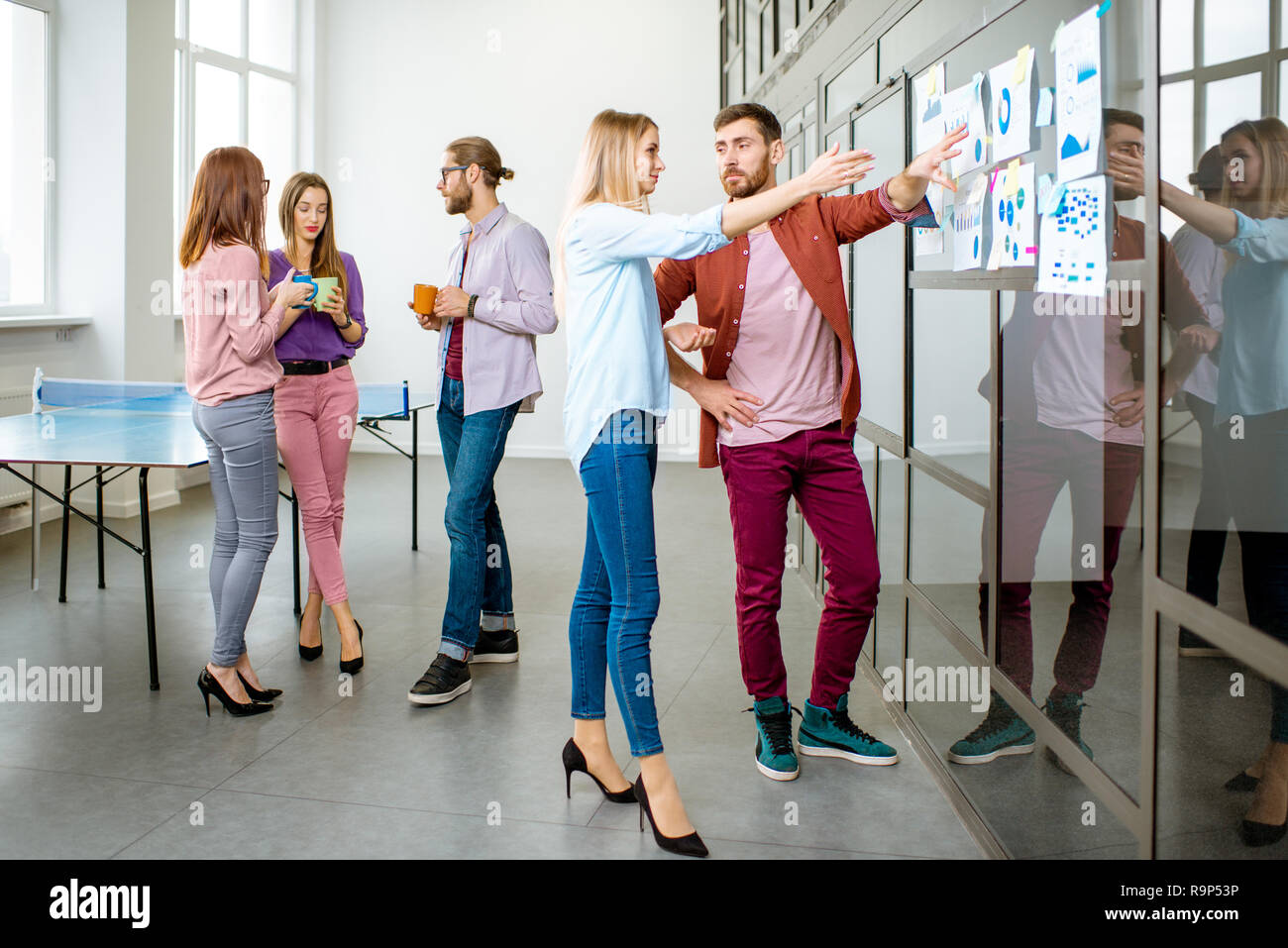 Junges Team von Mitarbeitern gekleidet beiläufig zusammen gesprochen in der Nähe der Glaswand mit einige Statistiken und Aufkleber im Büro Stockfoto