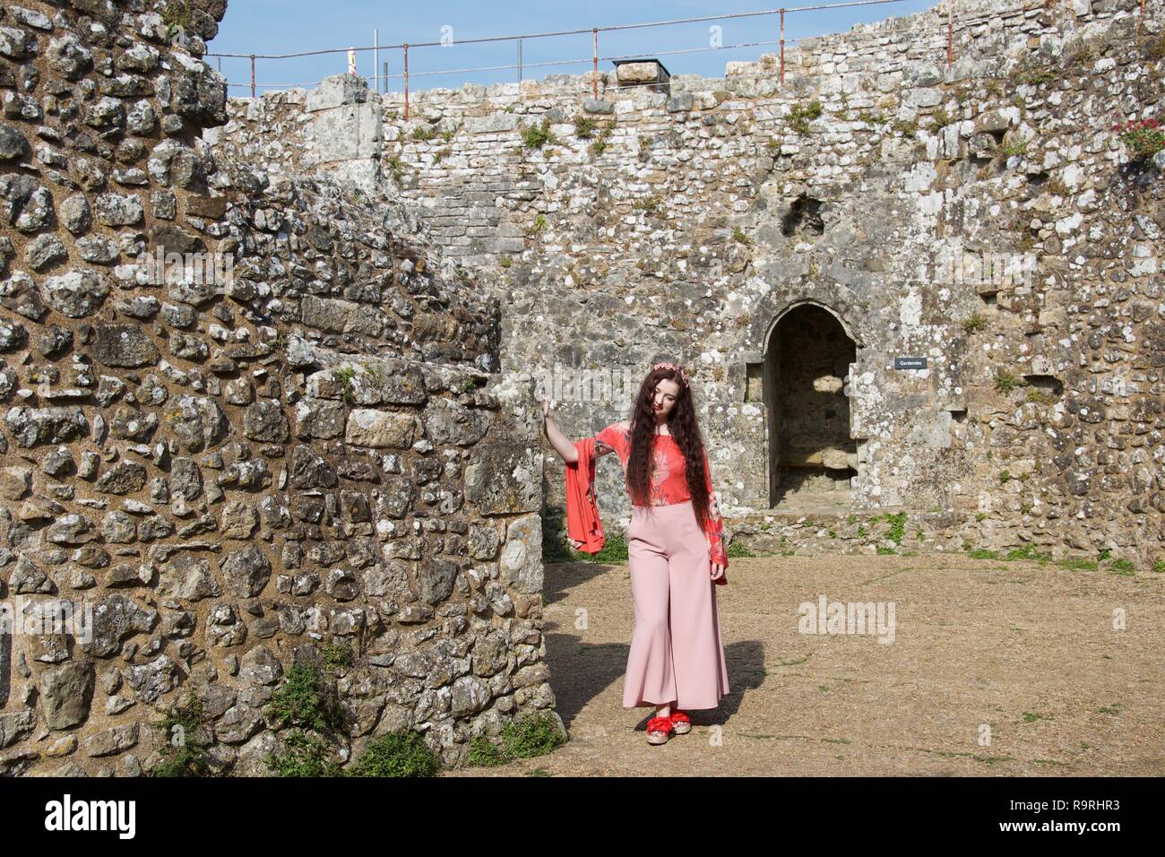 Ein hippie Lady mit rosa Schlaghosen, Modisch weite Ärmel und Haare Blumen, posees in der Sonne, gegen die Steinmauer in einem zerstörten Engl schiefen Stockfoto