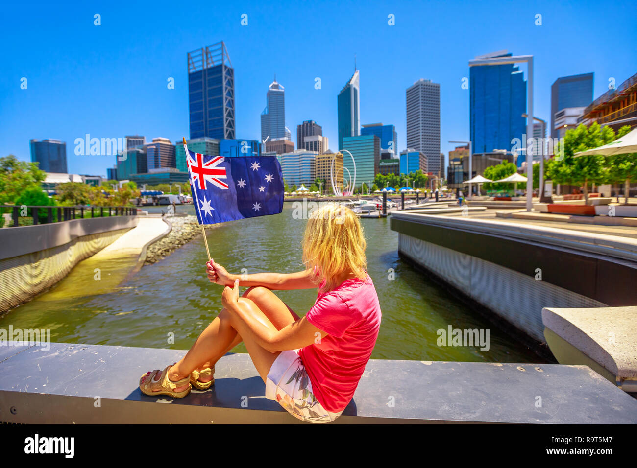Blonde Frau mit der australischen Flagge auf Elizabeth Quay Marina Promenade an einem sonnigen Tag. Central Business District in Perth, Western Australia auf verschwommenen Hintergrund. Kaukasische tourist genießt einen Blick auf die Stadt. Stockfoto