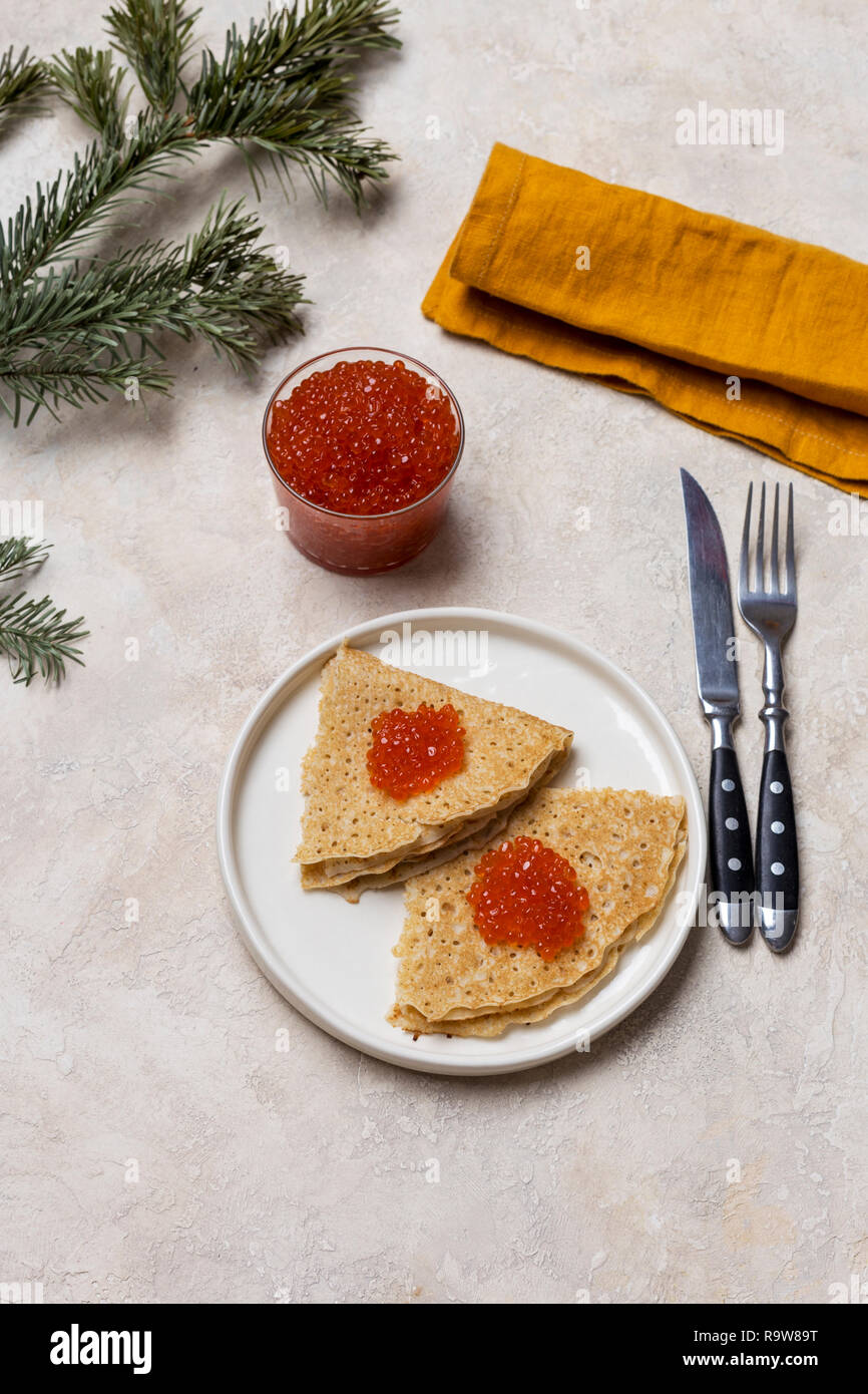 Dünne Pfannkuchen mit roter Kaviar an weiße Platte mit Glas mit rotem Kaviar, Gabel, Messer, Serviette und Tanne in der Nähe von hellen Hintergrund Stockfoto