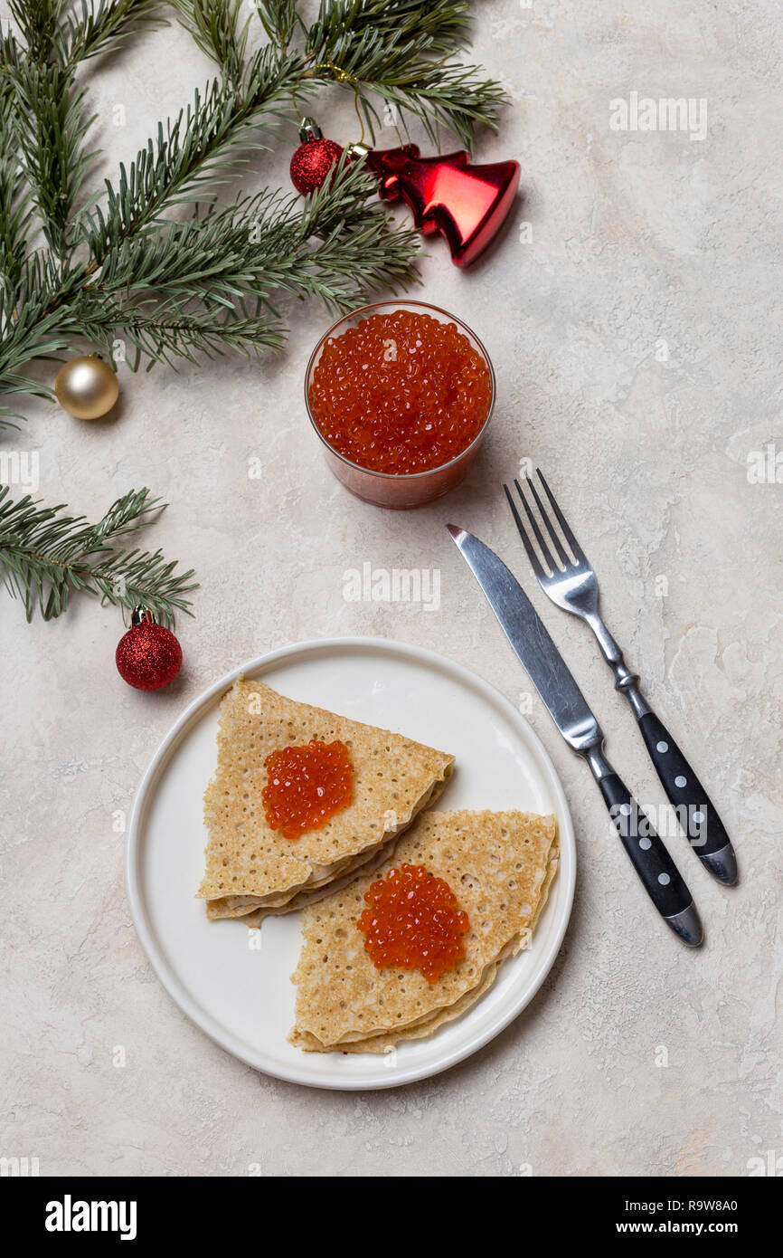 Das neue Jahr Frühstück. Dünne leckere Pfannkuchen mit roter Kaviar an weiße Platte mit Glas mit rotem Kaviar, Gabel, Messer, Weihnachten Spielzeug und Tanne in der Nähe an der Bac Stockfoto