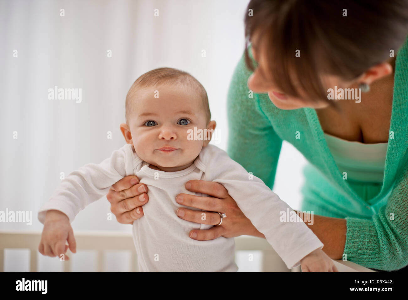Porträt eines lächelnden baby boy von den Händen seiner Mutter unterstützt werden, als sie ihm hilft, sich in seinem Kinderbett stehen. Stockfoto