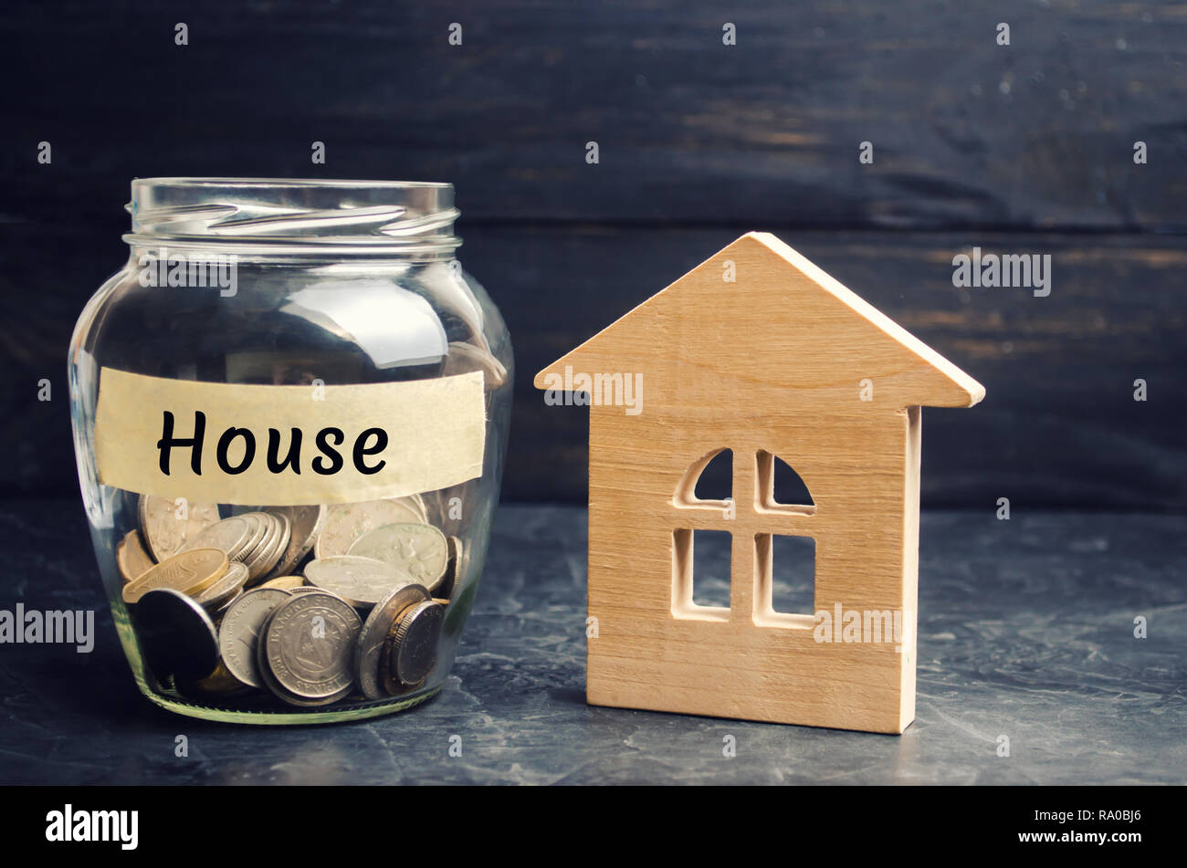Ein Glas Glas mit Münzen und ein Holzhaus und die Aufschrift 'Haus'. Die Anhäufung von Geld, um ein Haus zu kaufen. Mieten Sie eine Apartments. Kauf eines Hauses. Stockfoto