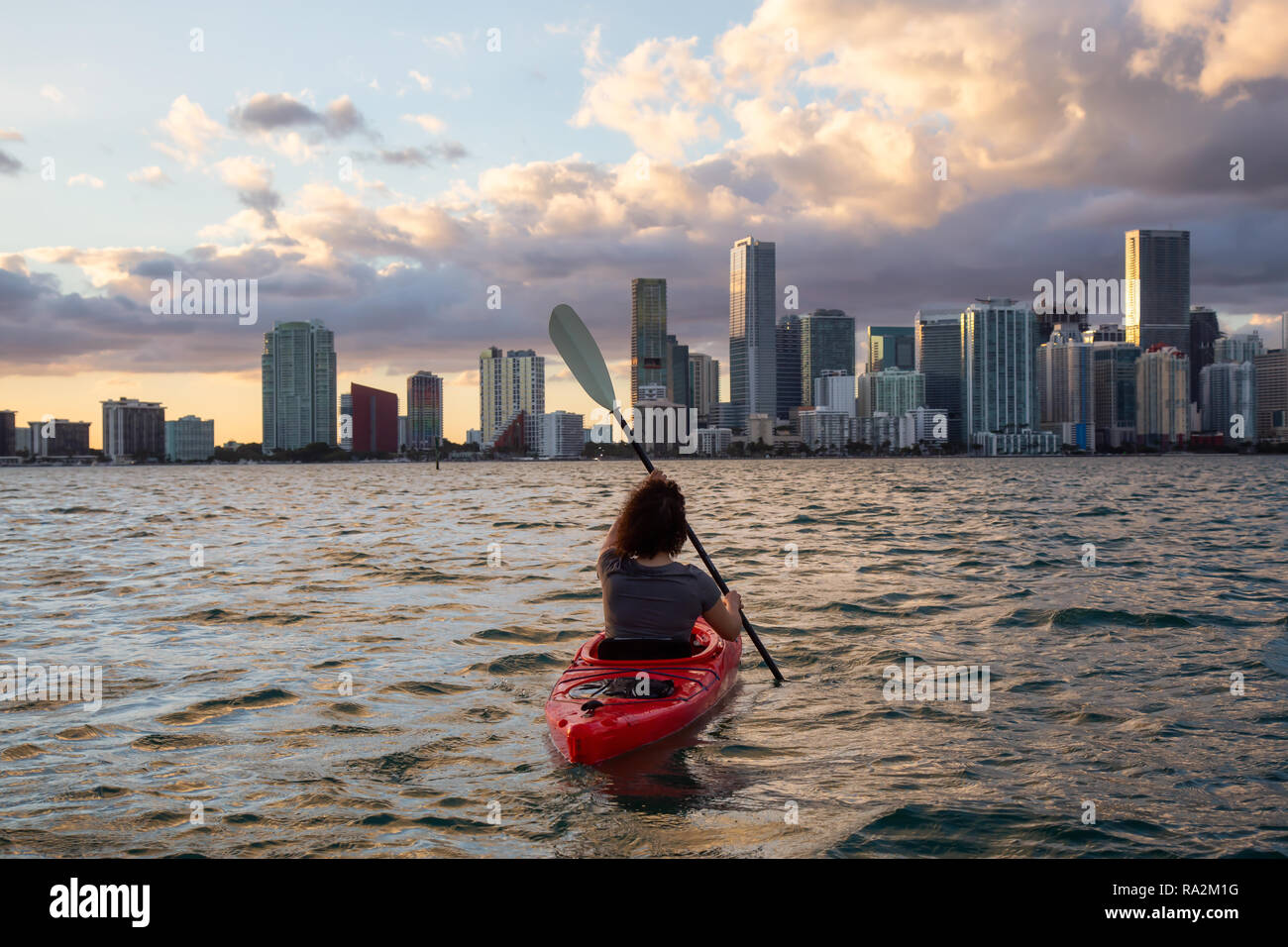 Abenteuerliche Mädchen Kajak vor einem modernen Stadtzentrum Stadtbild in einer dramatischen Sonnenuntergang. In Miami, Florida, Vereinigte Staaten von Amerika übernommen. Stockfoto