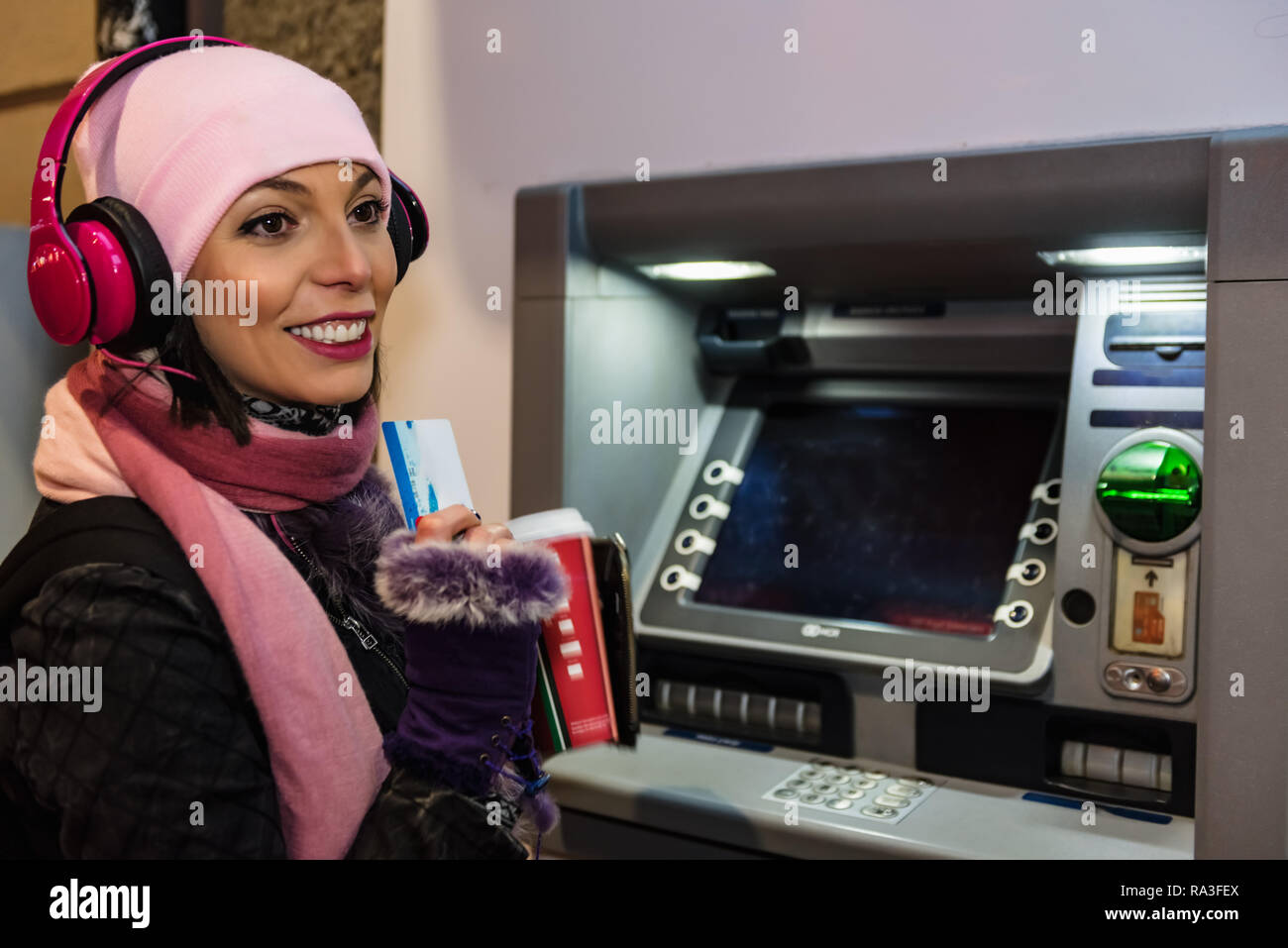 Schöne Frau in beanie mit Kaffee zum Mitnehmen und Kopfhörer zieht in ec-Karte an einem Geldautomaten. ATM cash-Terminal mit Display Stockfoto