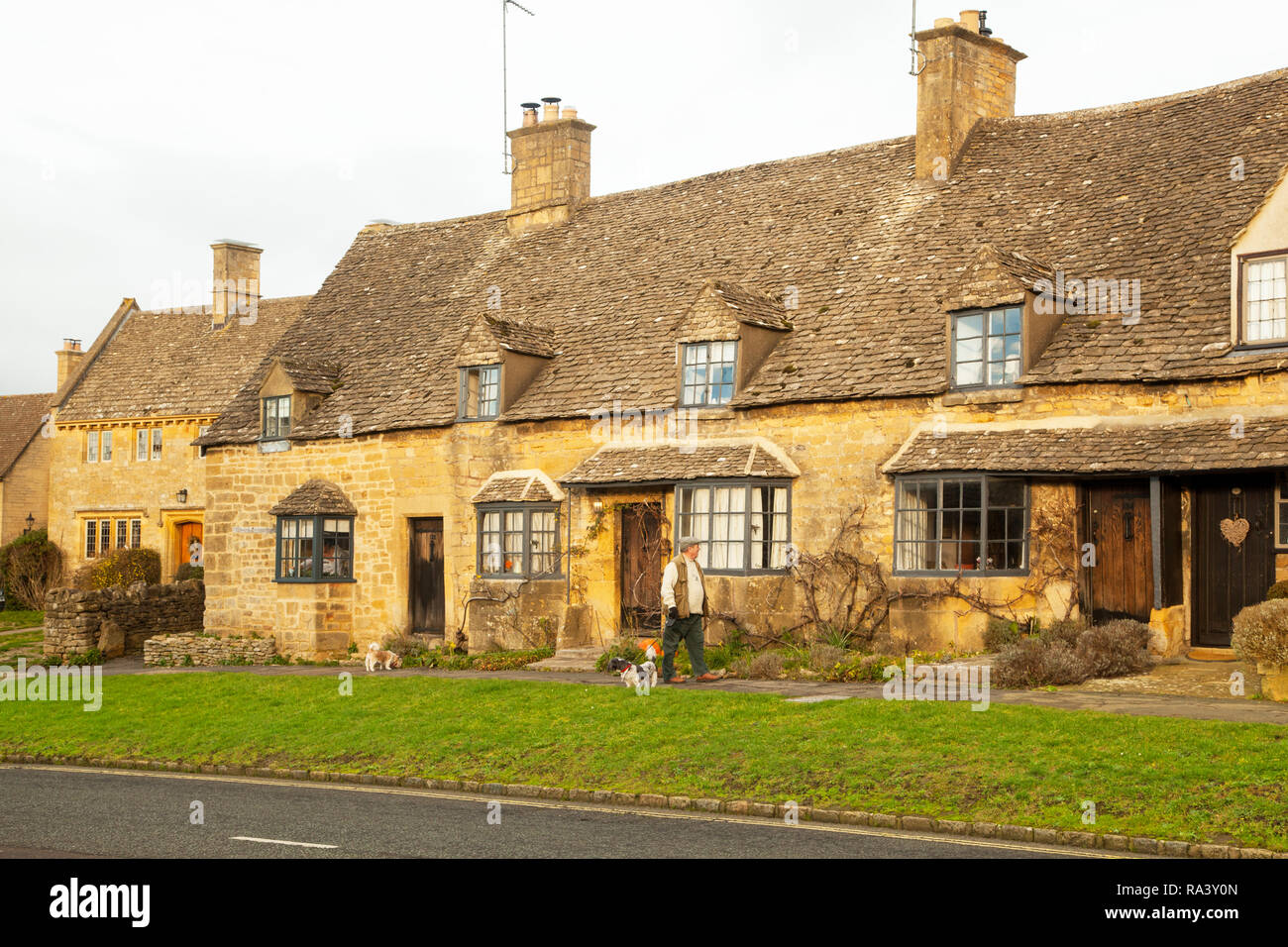 Mann, seine Hunde durch das malerische Dorf Cotswold schlechthin der Broadway mit seinen Cotswold stone Gebäude und Häuser Stockfoto