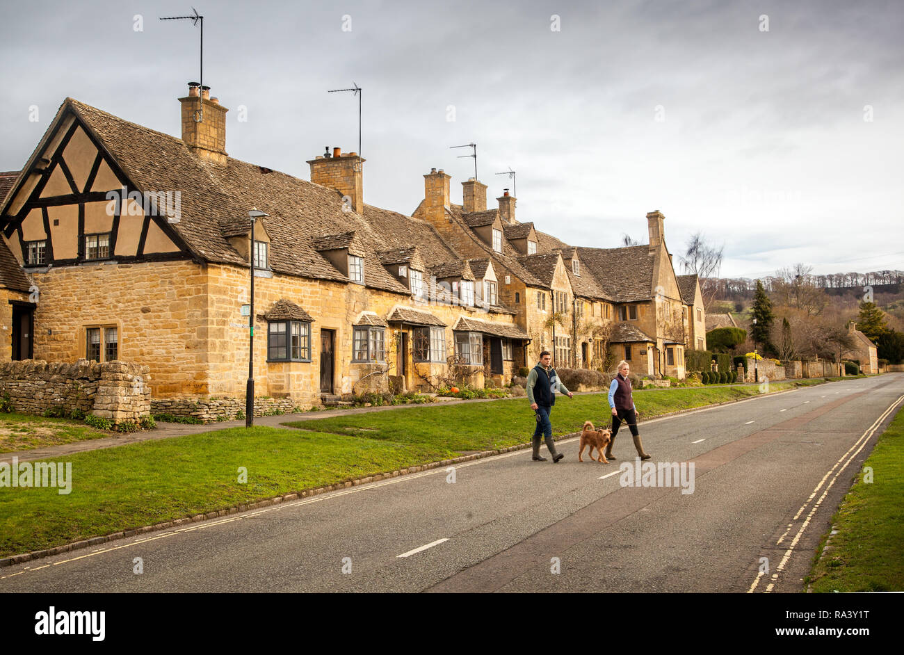 Der Mann und die Frau zu Fuß einen Hund durch das malerische Dorf Cotswold schlechthin der Broadway mit seinen Cotswold stone Gebäude und Häuser Stockfoto