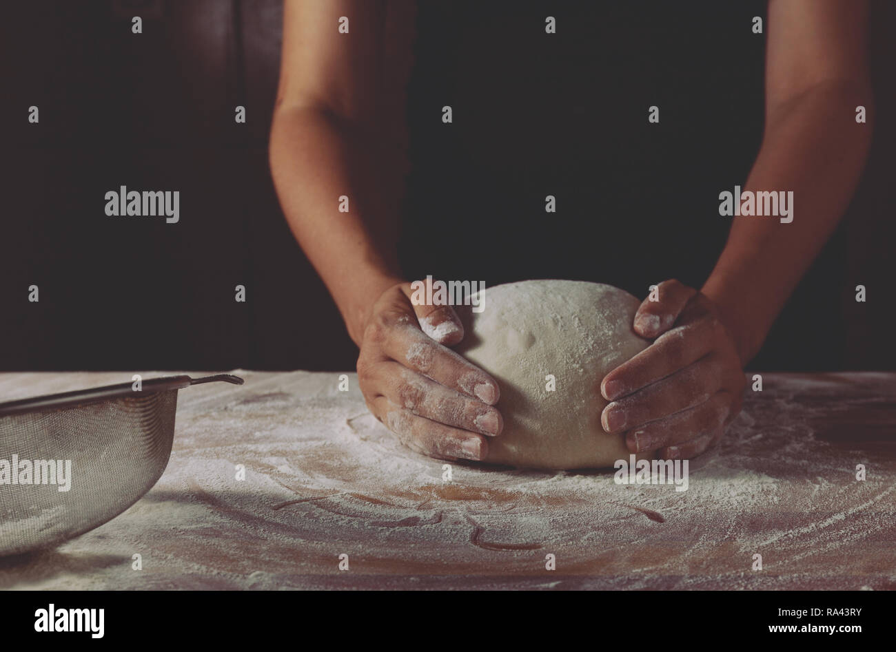 Der Teig durch weibliche Hände auf einer hölzernen backen Tabelle. Dunkler schwarzer Hintergrund mit Mehl. Seitenansicht, horizontale Foto Stockfoto
