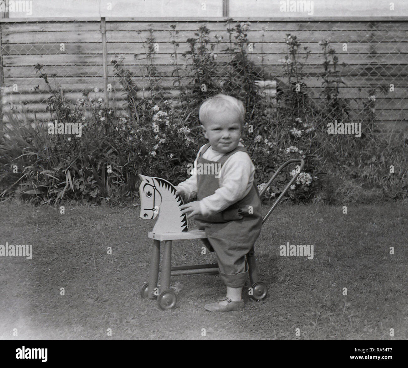 1960, historische, ein Kleinkind junge außerhalb in einem Garten auf einer hölzernen Spielzeug Pferd mit kleinen Rädern. Stockfoto