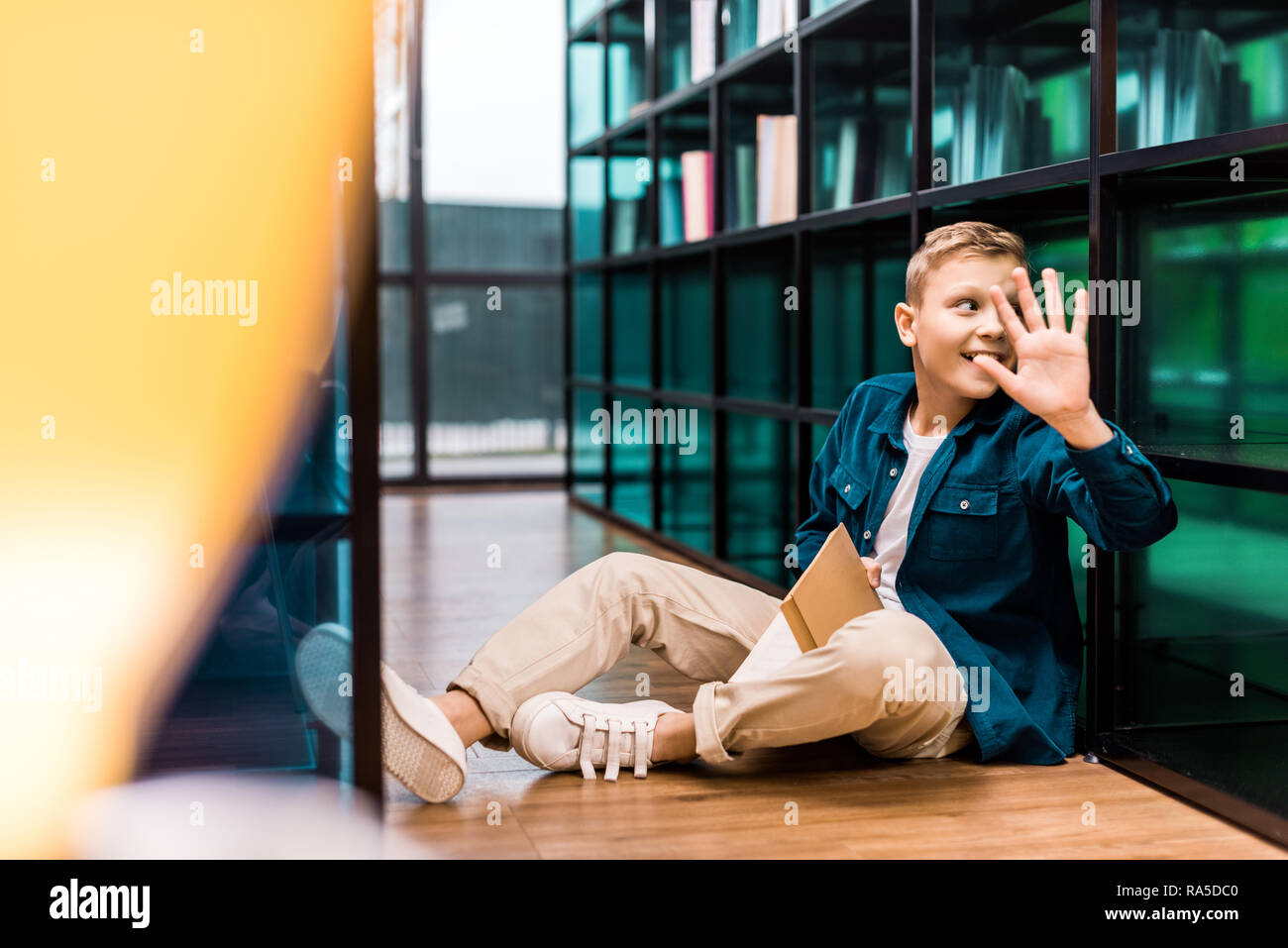 Lächelnden Schuljungen holding Buch und winkende Hand beim Sitzen auf dem Boden in der Bibliothek Stockfoto