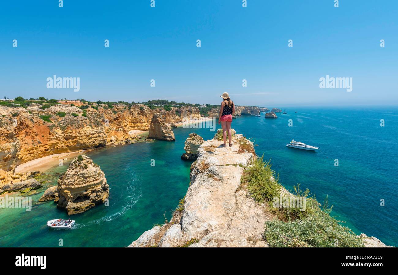 Junge Frau, die auf den Felsen an der Steilküste, mit Blick auf das türkisfarbene Meer, Strand Praia da Marinha, zerklüftete Felsküste aus Sandstein Stockfoto
