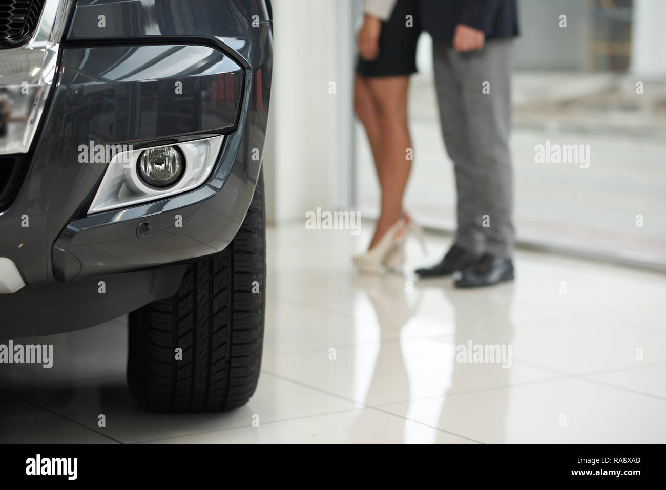 Scheinwerfer und Reifen von teuren schwarzen Auto. Frau das Tragen von High Heels, Rock und Mann hinter stehen. Nicht wiederzuerkennen, Kunden, Auto im Autohaus. Stockfoto