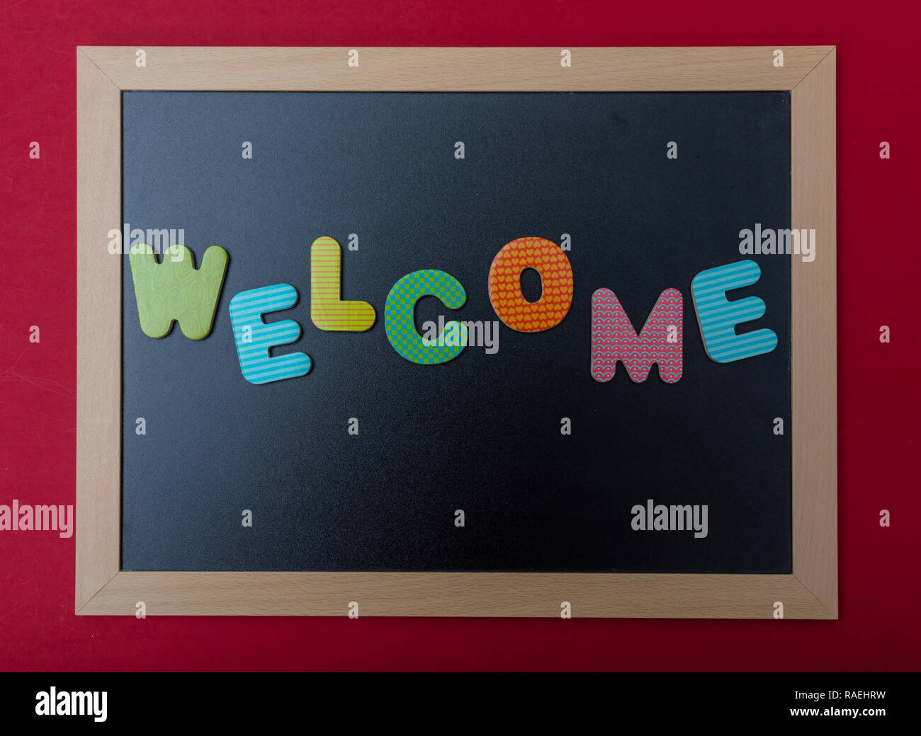 Willkommen. Vorstand mit schwarzem Rahmen, Text Willkommen in bunten Buchstaben, rote Wand Hintergrund Stockfoto